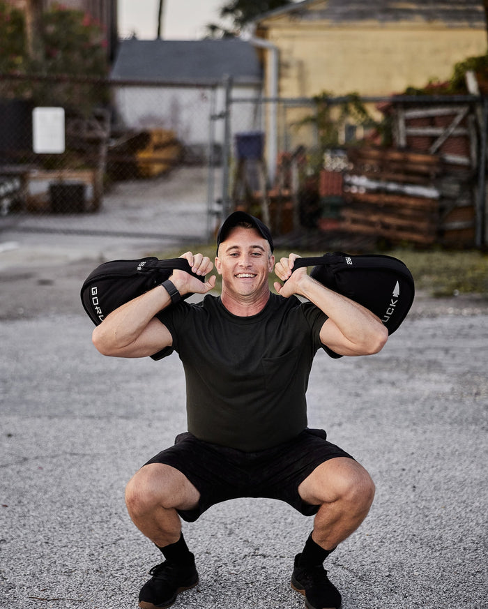 Man performing outdoor squat with GORUCK sandbags, wearing athletic gear, urban background