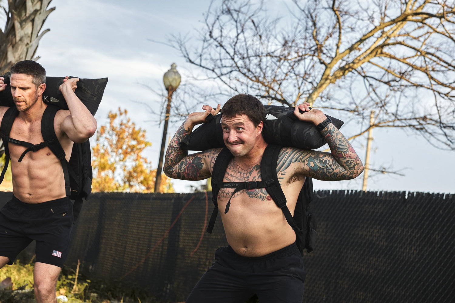 Two men with rucksacks and sandbags exercise outdoors, GORUCK fitness training scene