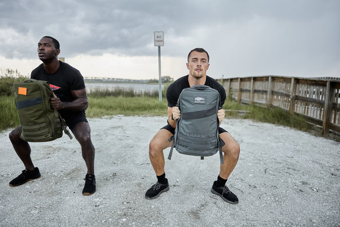 Two men doing squats outdoors with GORUCK rucksacks by a gravel path and wooden fence