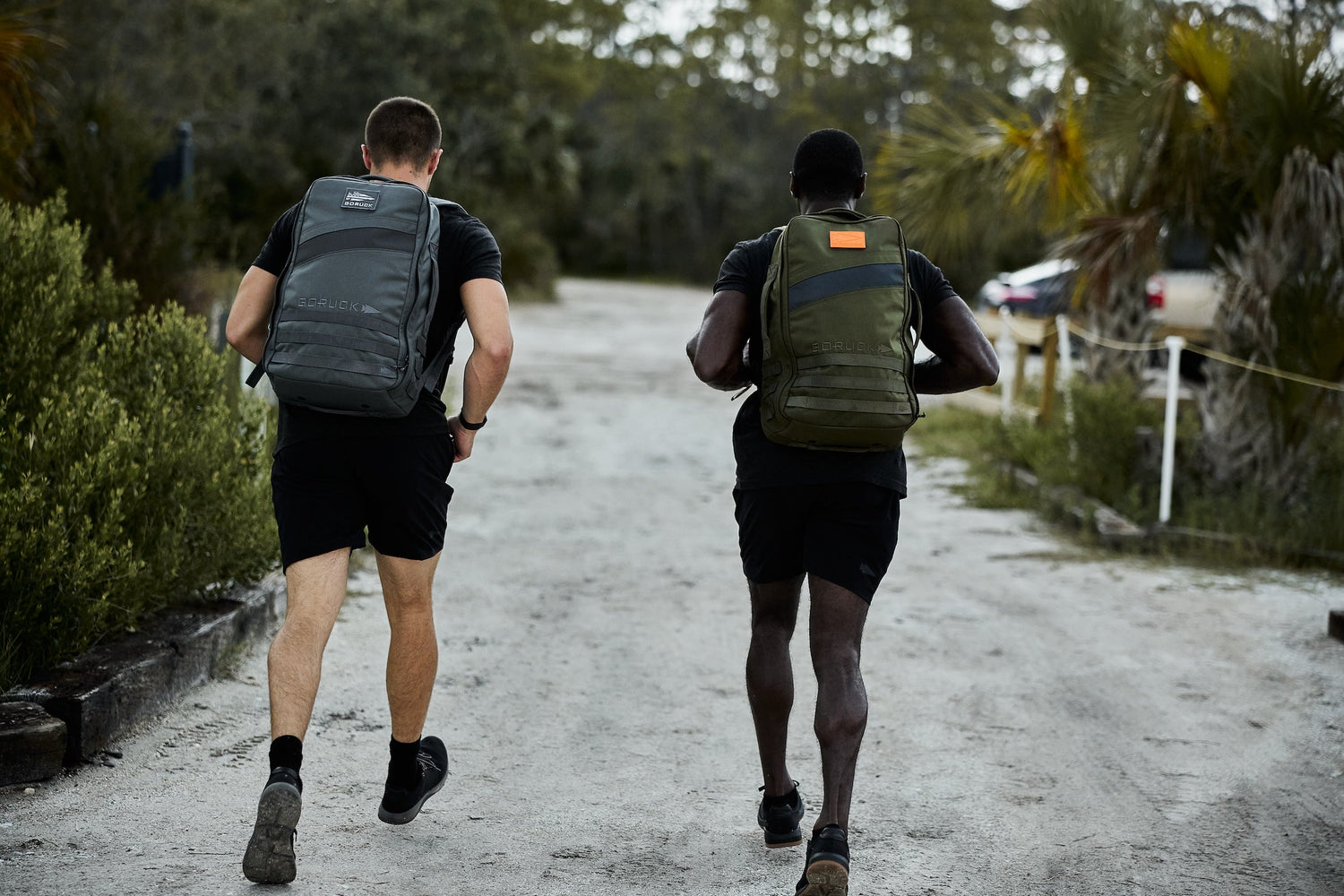 Two men wearing GORUCK backpacks rucking outdoors on a dirt path surrounded by greenery