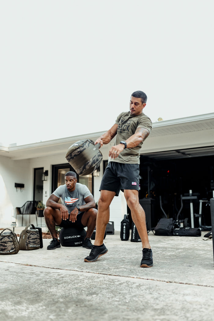 Two men training outdoors with GORUCK gear, one swinging a sandbag, rugged fitness setting.