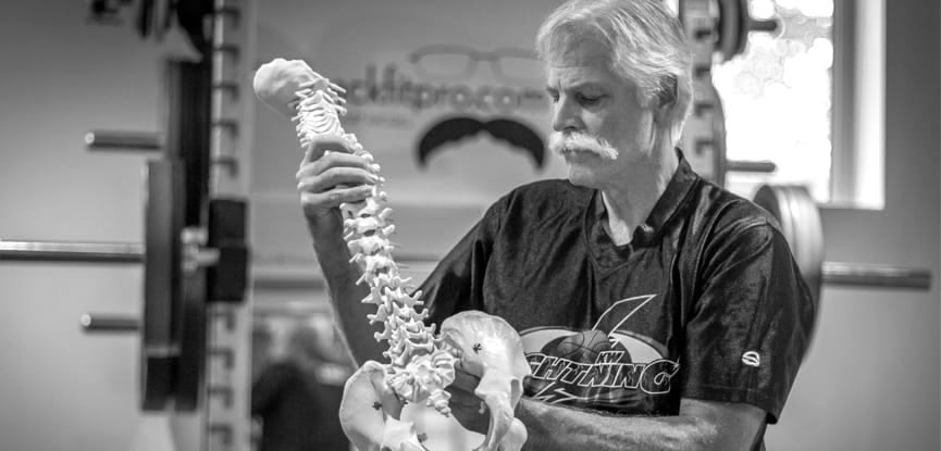 Older man in gym holding anatomical spine model, strength training equipment in background, GORUCK focus