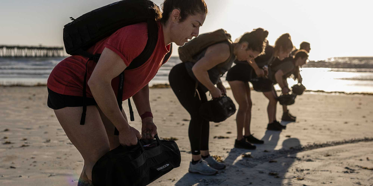 Group workout with GORUCK gear on beach, people training with rucksacks and sandbags