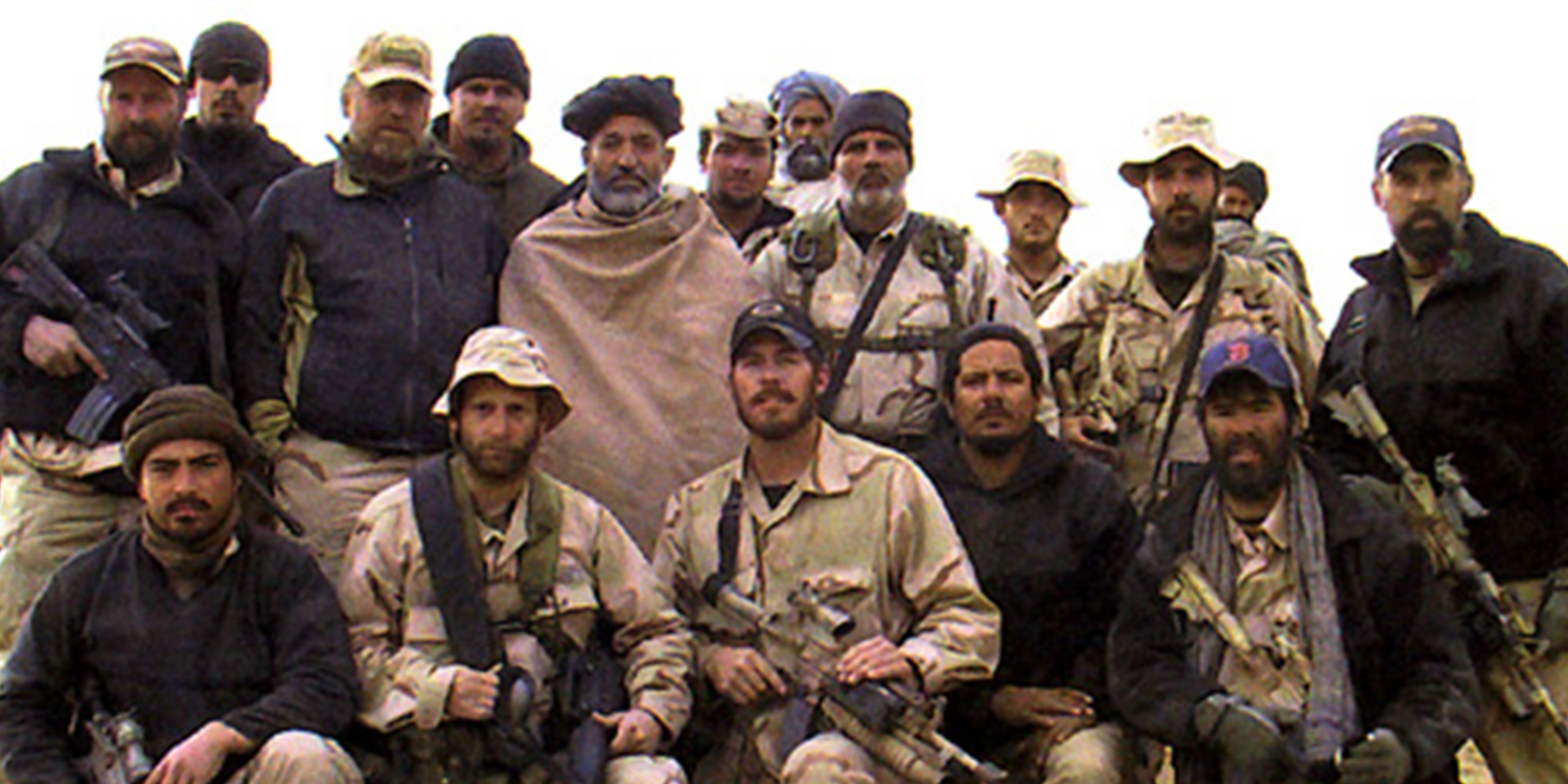 Group of men in tactical and outdoor gear posing outdoors, some holding rucking equipment, GORUCK style.