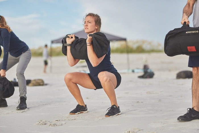 Woman doing sandbag squat on beach during GORUCK outdoor workout event