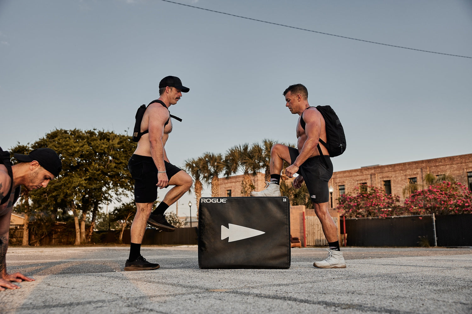 Shirtless men rucking with GORUCK backpacks doing outdoor fitness workouts by a Rogue plyo box
