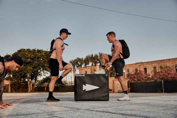 Shirtless men rucking with GORUCK backpacks doing outdoor fitness workouts by a Rogue plyo box