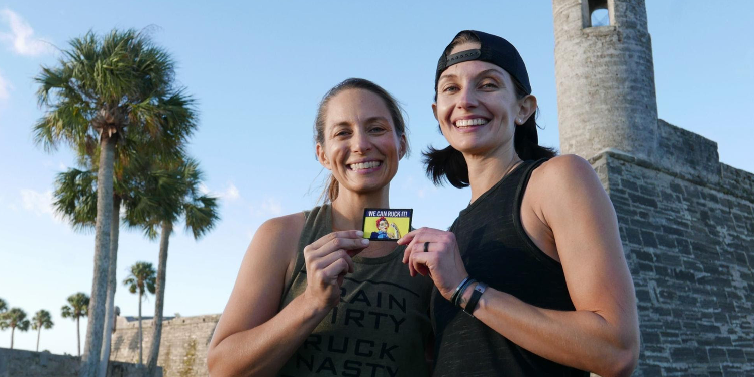 Two women smiling and holding 'We Can Ruck It!' patch, palm trees and stone tower in background, GORUCK event