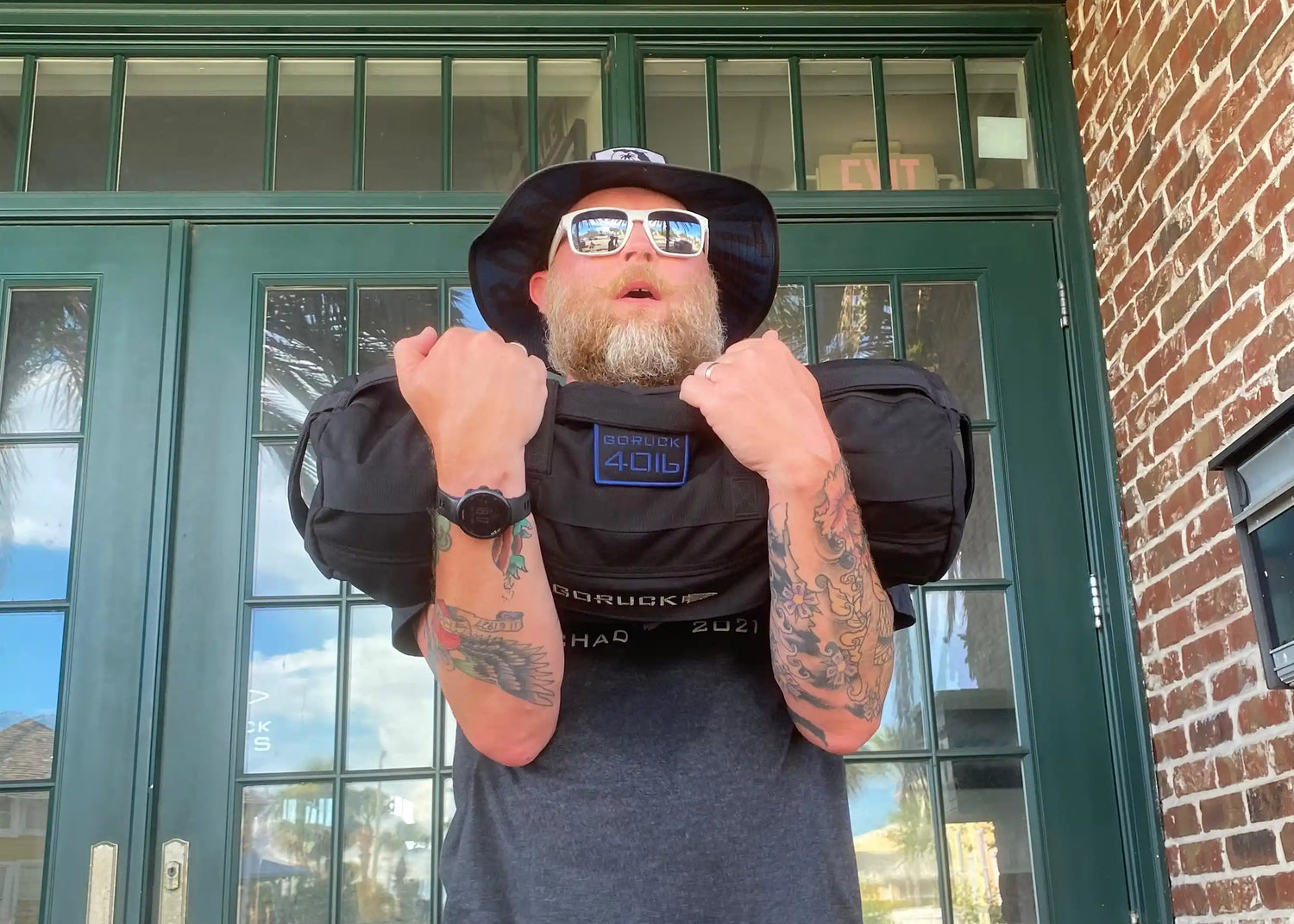 Bearded man with tattoos lifting GORUCK weighted sandbag outdoors, showing tough rucking gear.