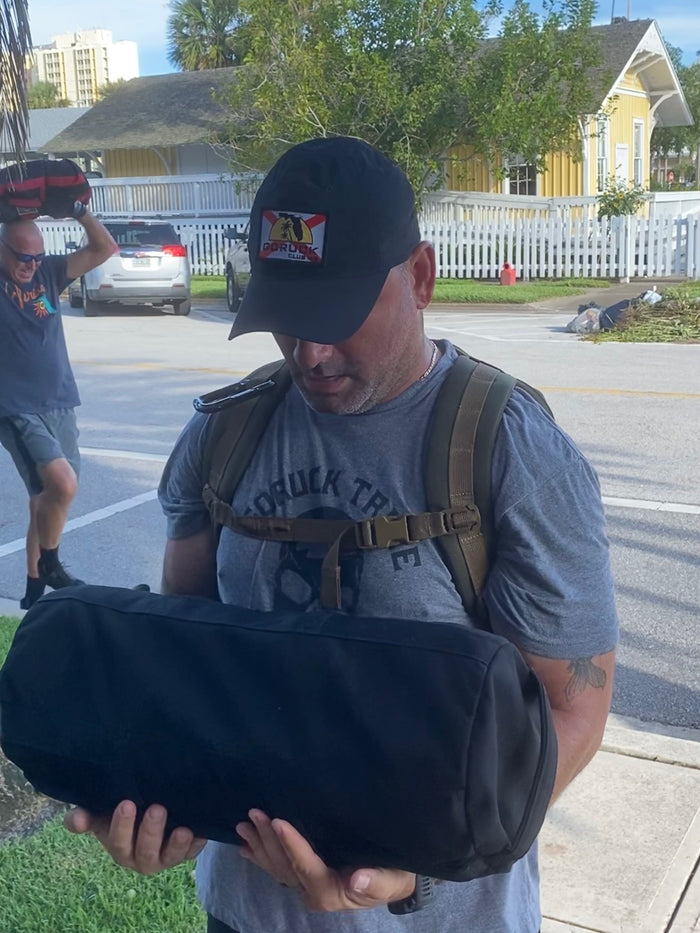 Man in GORUCK gear carrying weighted sandbag outdoors, participating in rucking event