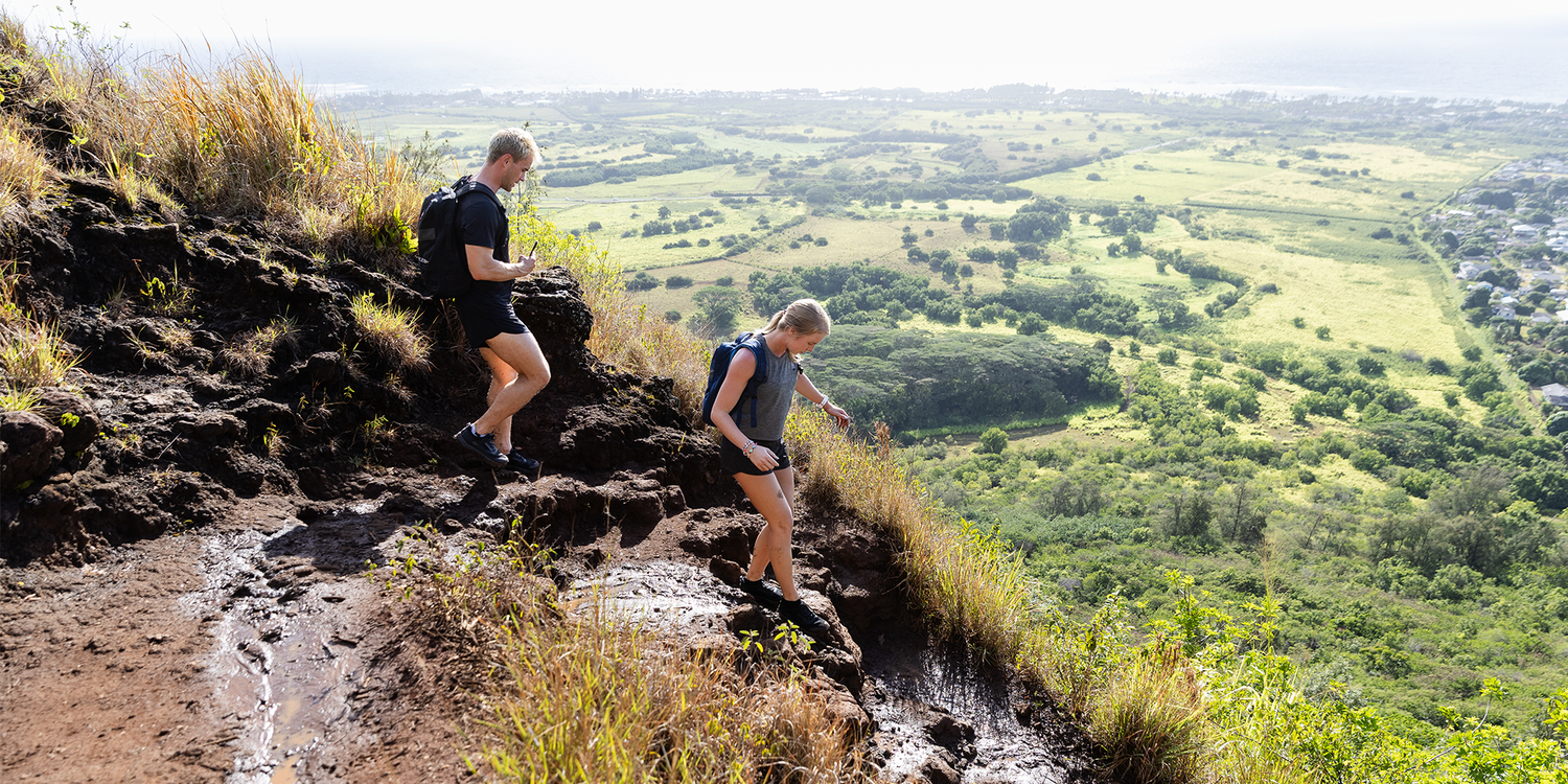 Two hikers in GORUCK gear trekking a muddy trail on a hill with a scenic valley view