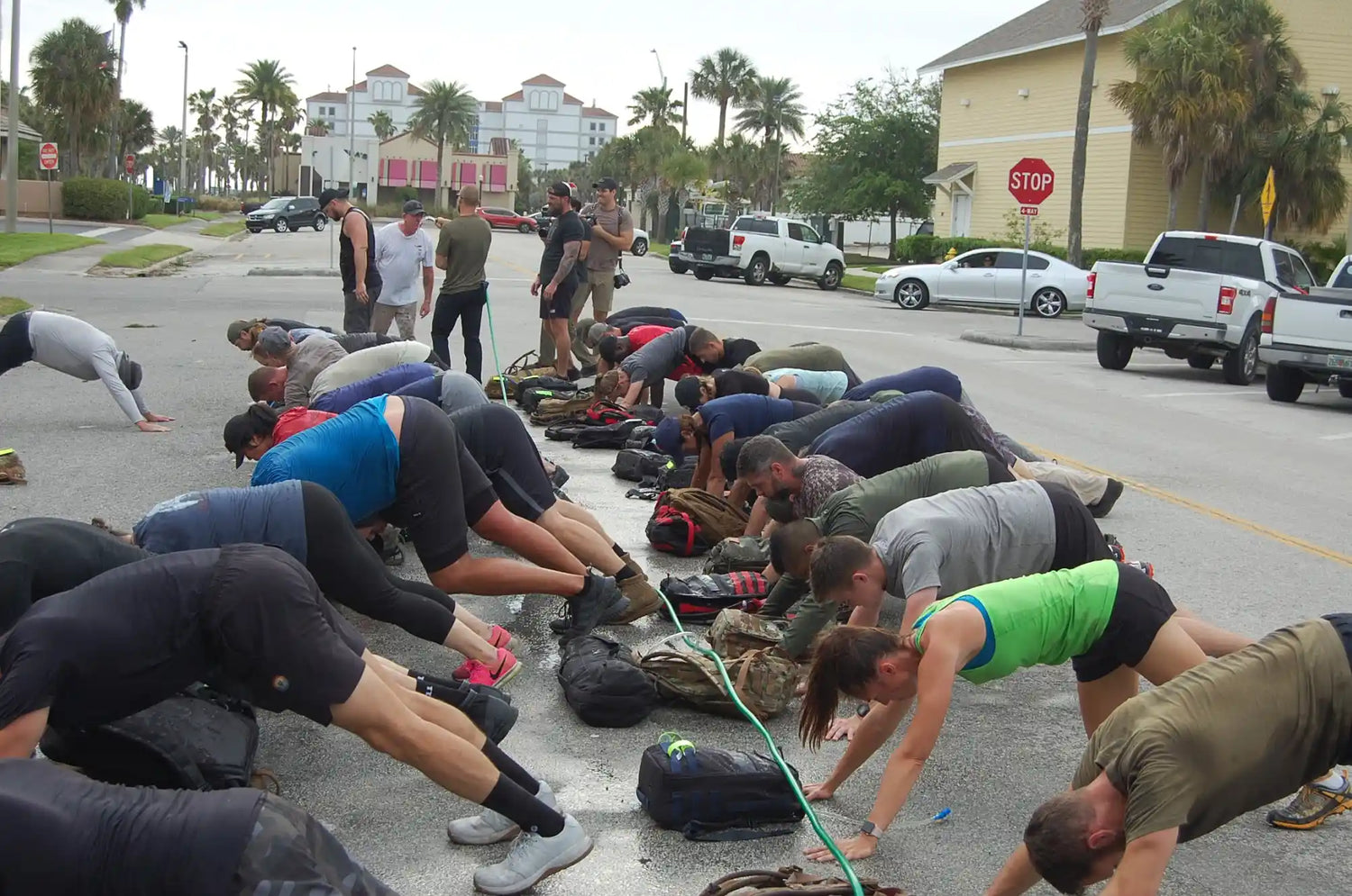 Outdoor GORUCK fitness group doing push-ups on asphalt with backpacks and palm trees in background
