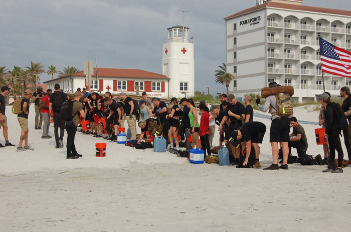 GORUCK rucking event participants on beach with gear, buckets, lighthouse, and American flag visible