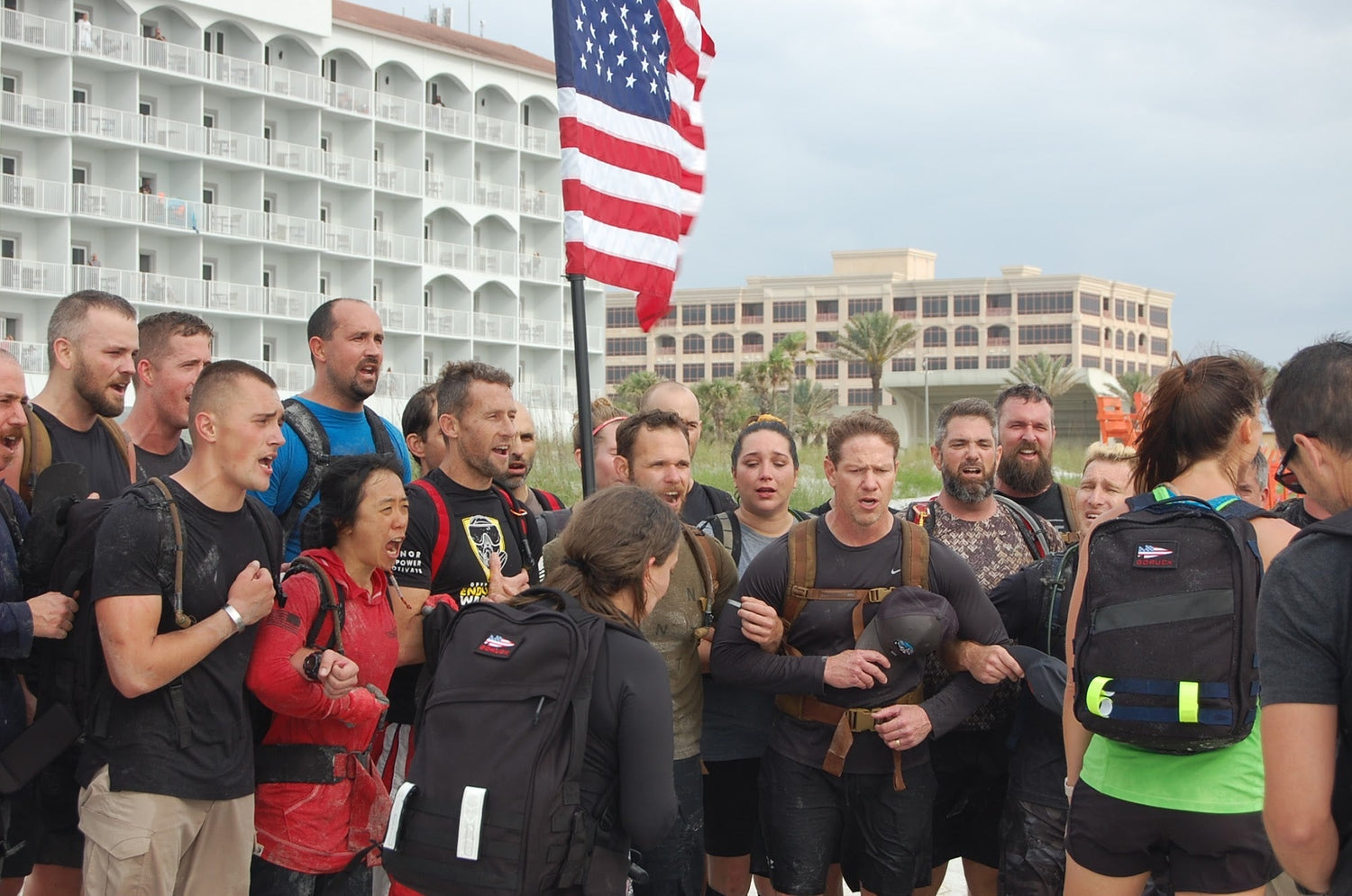 GORUCK participants with rucksacks standing on a beach, holding an American flag, group event