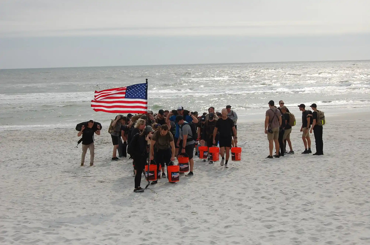 GORUCK group rucking on sandy beach with American flag and training buckets