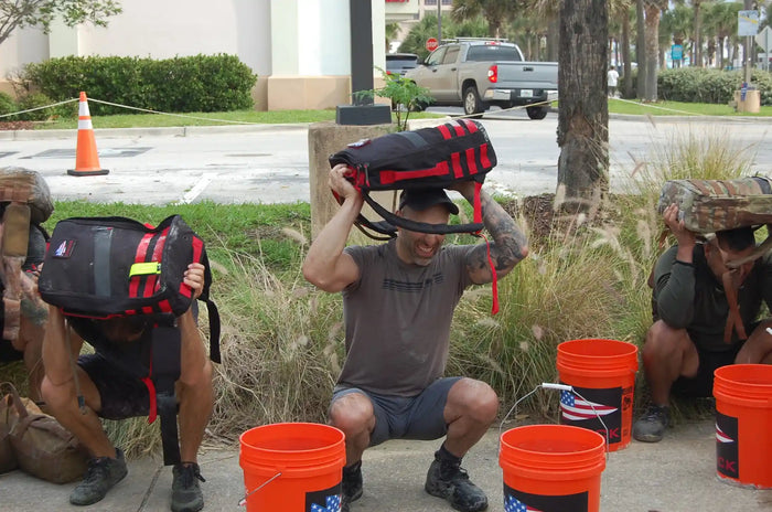 Three people outdoors squatting with GORUCK rucksacks overhead near orange buckets during a rucking fitness event