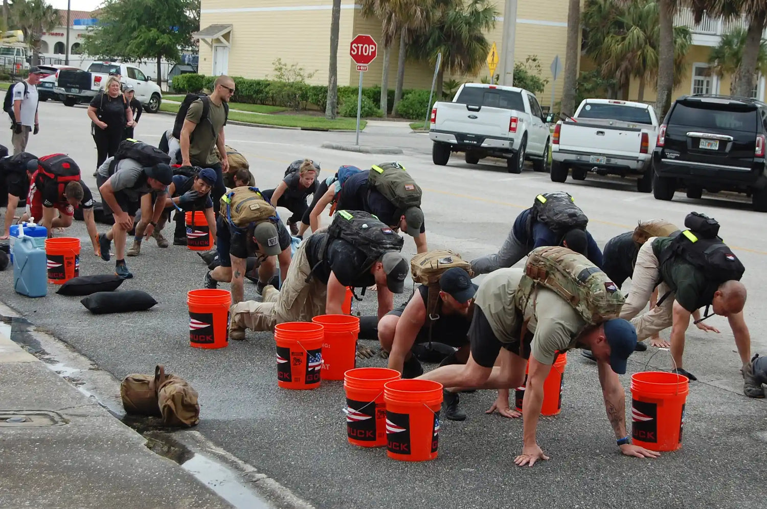 GORUCK rucking event participants wearing backpacks perform bear crawls outdoors next to orange GORUCK buckets.