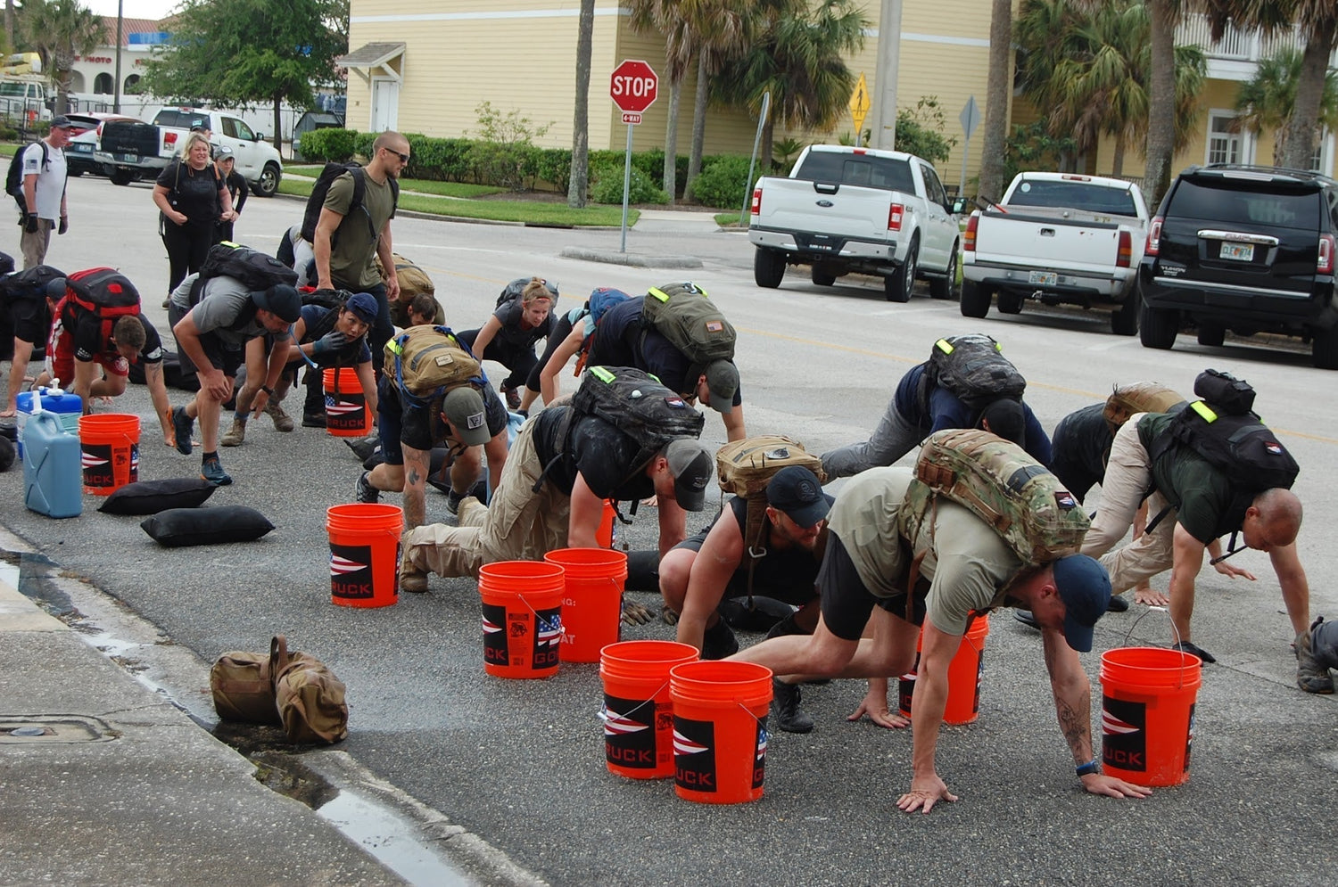 GORUCK participants with rucksacks doing outdoor team training drills on pavement with orange buckets