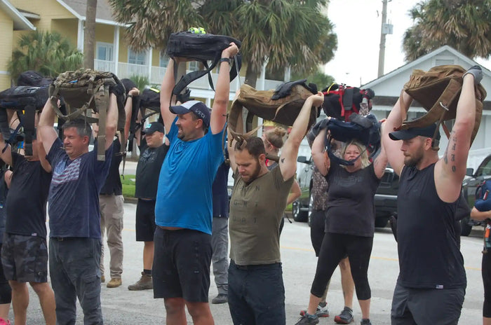 Group rucking workout with GORUCK gear, participants lifting rucksacks overhead outdoors