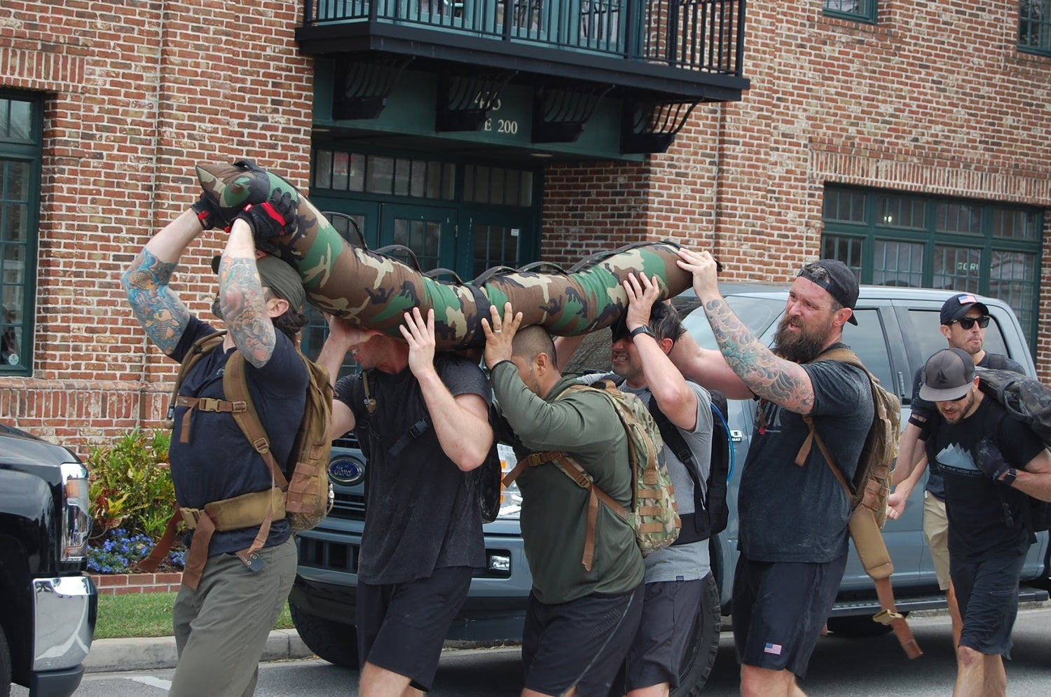 GORUCK participants team-carry heavy camo sandbag outdoors in rucking event, wearing gear
