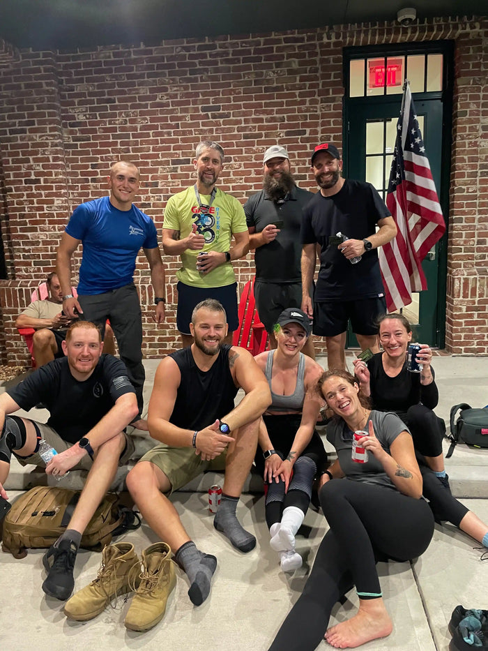 Smiling GORUCK rucking team relaxing indoors, posing with an American flag and gear bags.