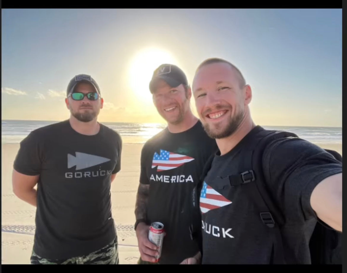 Three men in GORUCK shirts on a beach at sunrise, smiling and ready for rucking.