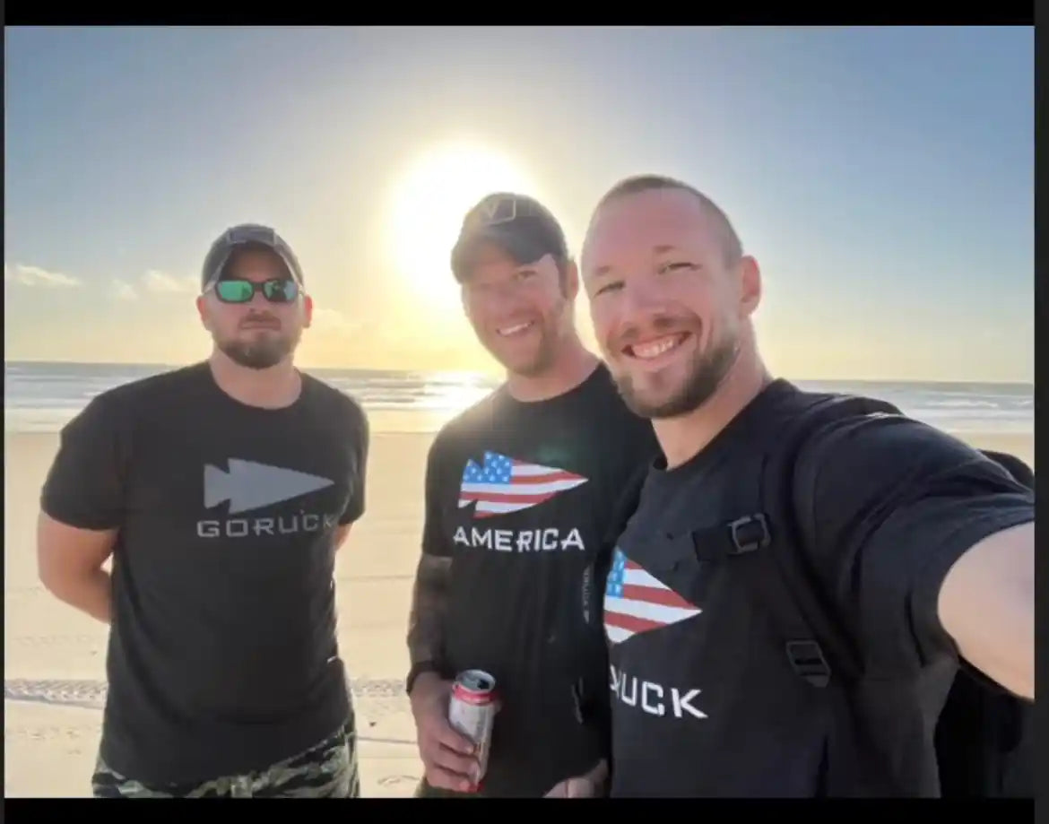 Three men in GORUCK shirts smiling on a sunny beach, representing rucking community.