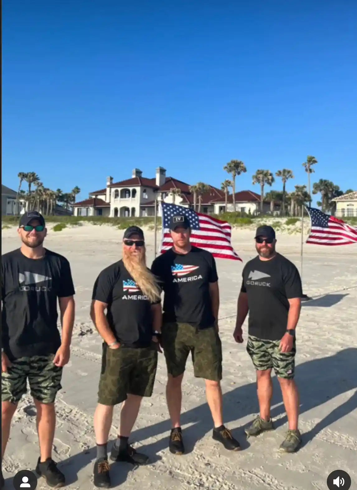 Four men in GORUCK gear stand on a sandy beach with American flags and palm trees.