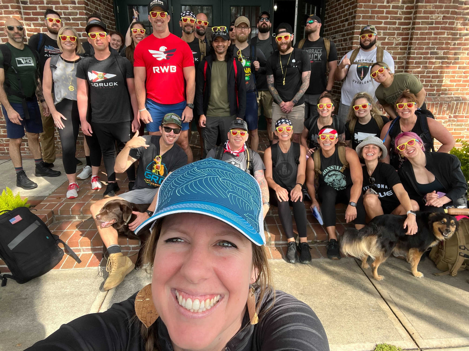 GORUCK group smiling outdoors with backpacks, sunglasses, and dogs at brick building entrance