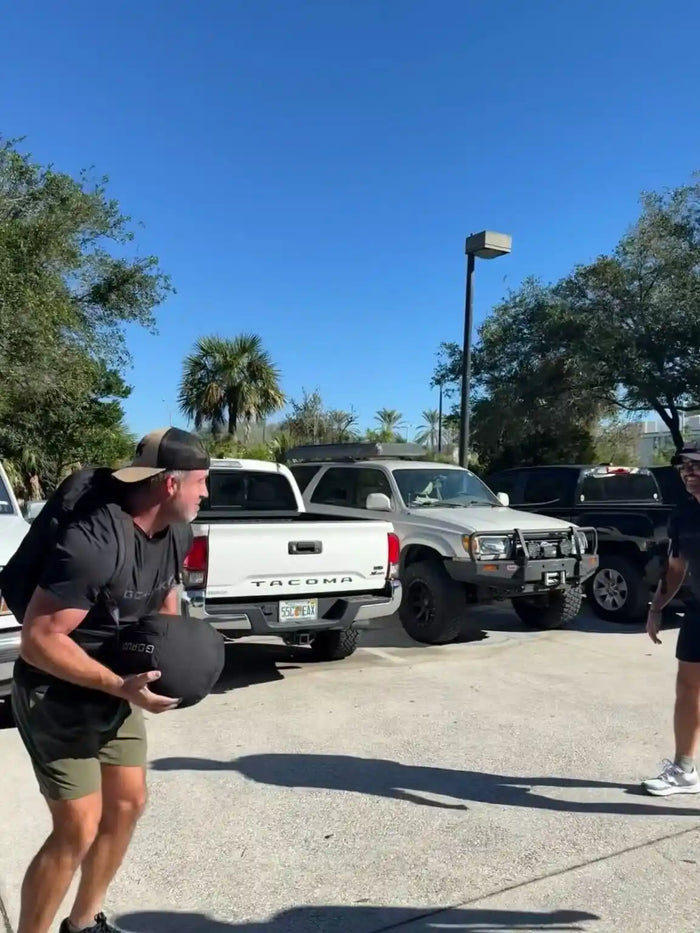 Man with GORUCK gear lifting sandbag outdoors in parking lot with palm trees
