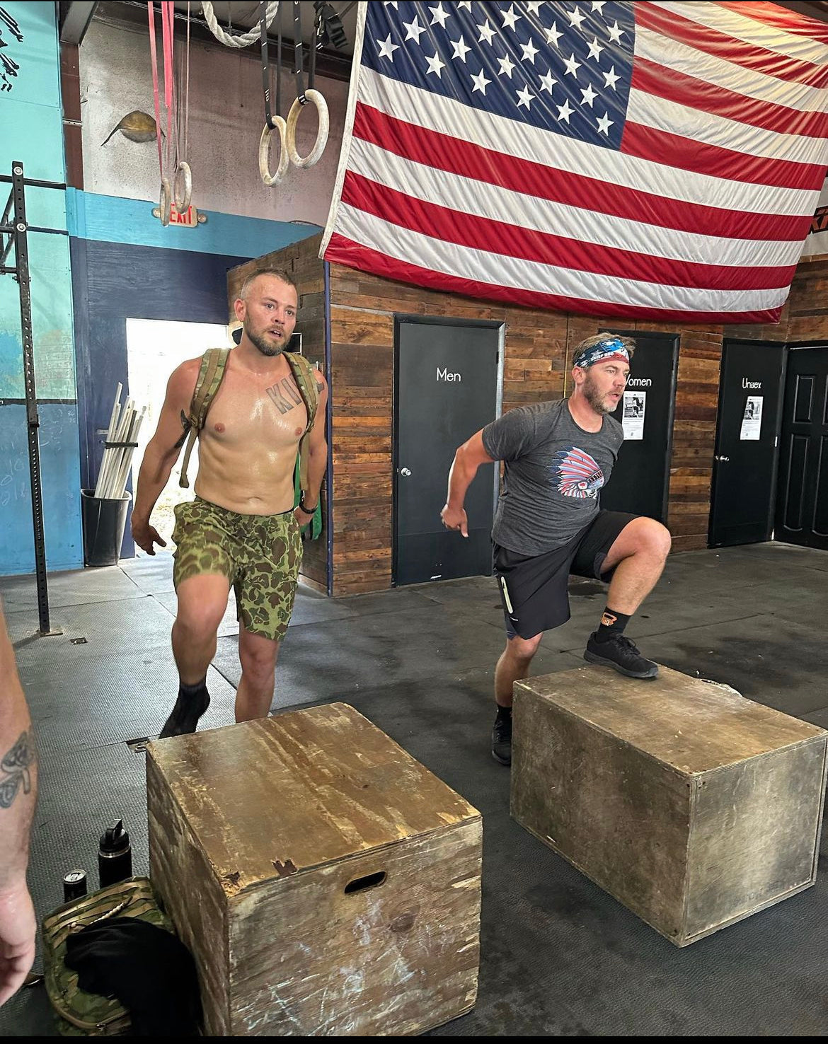 Two men perform box step-ups in a gym with an American flag, promoting GORUCK rucking gear.