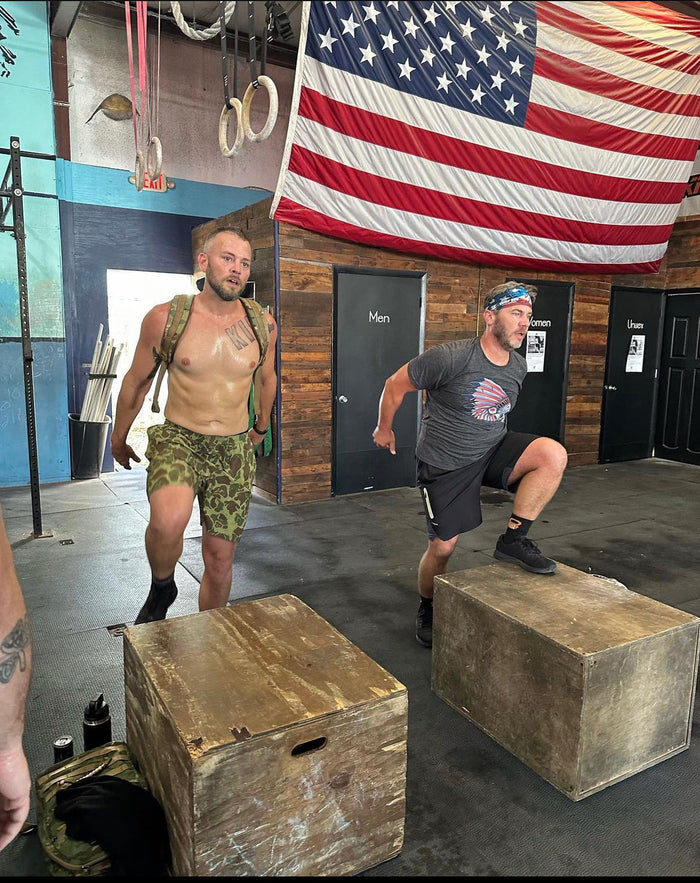 Two men perform box step-ups in a gym with an American flag, promoting GORUCK rucking gear.