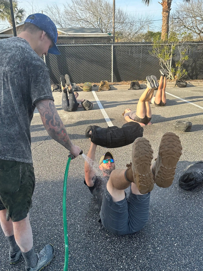 GORUCK outdoor fitness training, participants doing exercises with rucksacks, water hose spray