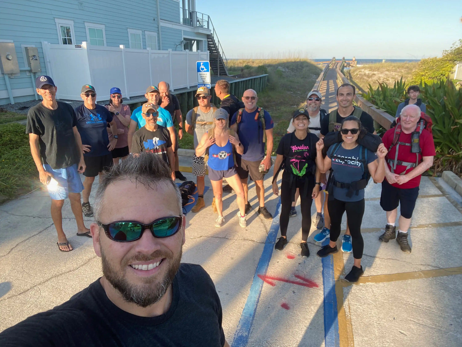 GORUCK group wearing rucksacks smiling outdoors near a beach boardwalk, ready for rucking event