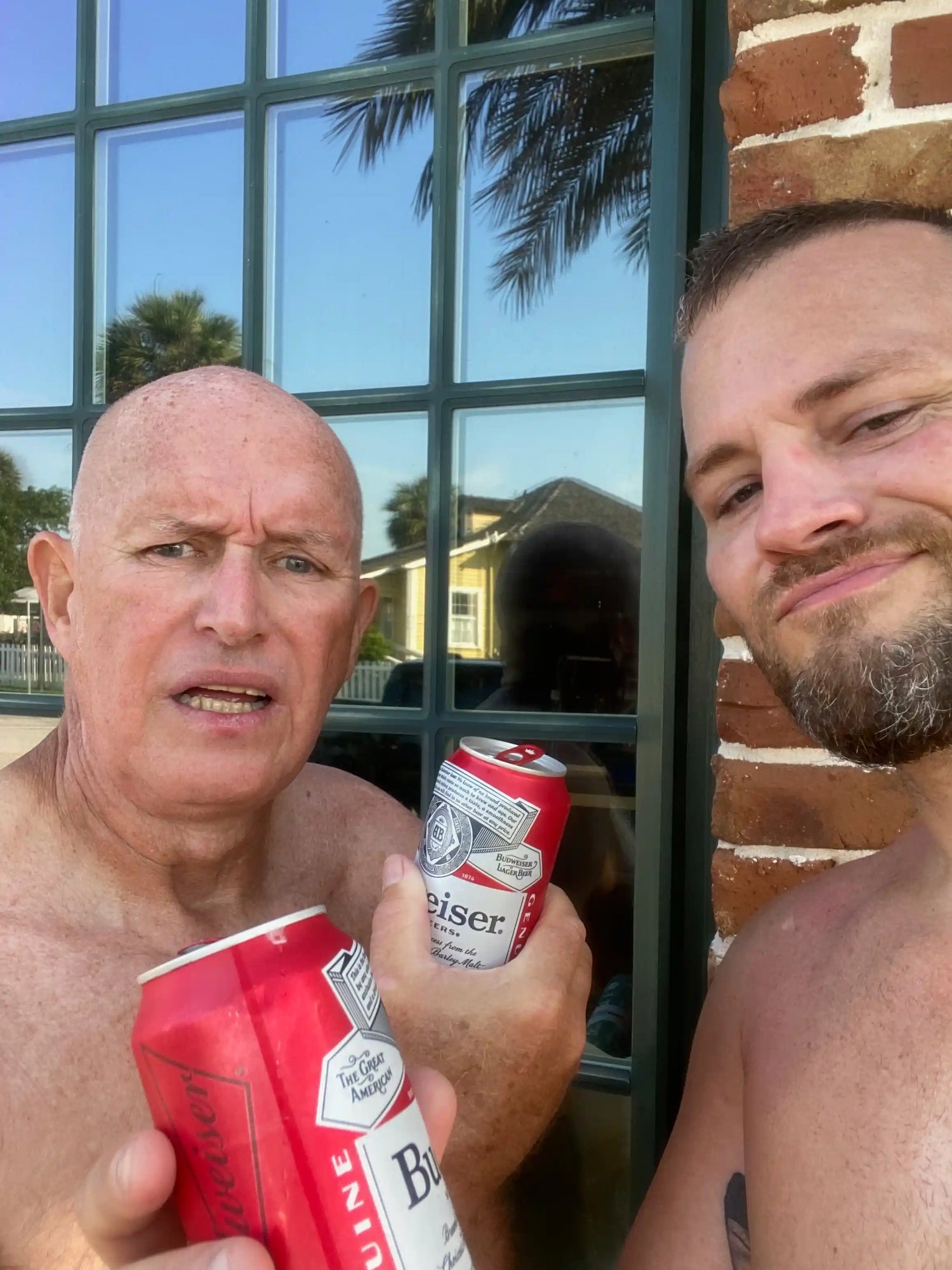 Two men outdoors holding Budweiser cans, standing by a window with palm tree reflections
