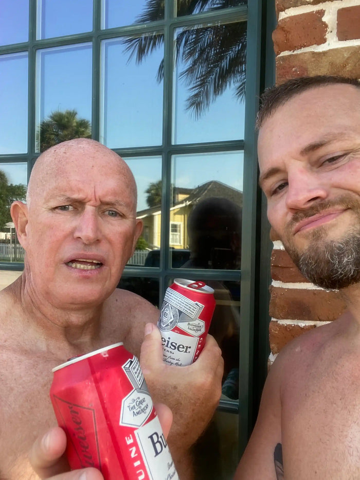 Two men outdoors holding Budweiser cans, standing by a window with palm tree reflections