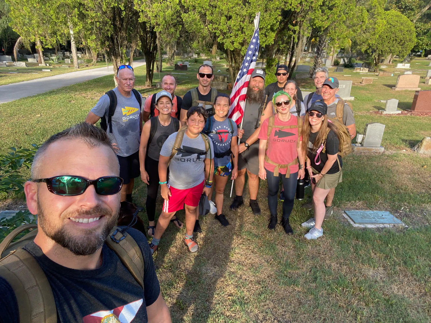 GORUCK group rucking outdoors with backpacks and American flag in a sunny cemetery