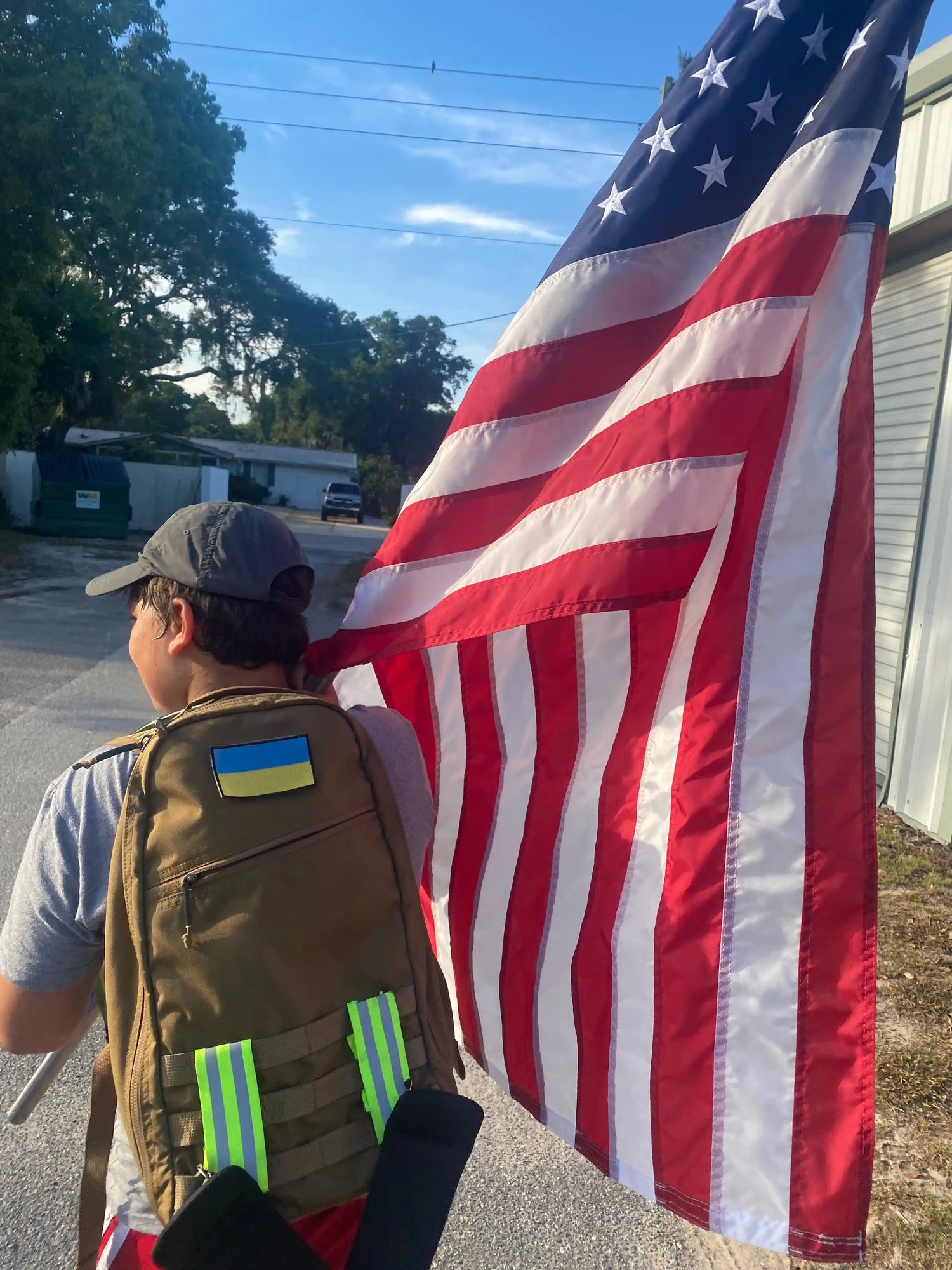 Person rucking with GORUCK backpack and large American flag outdoors on sunny day