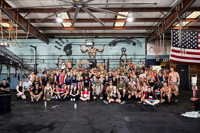 Large group of people at GORUCK fitness event in gym with wall art and American flag
