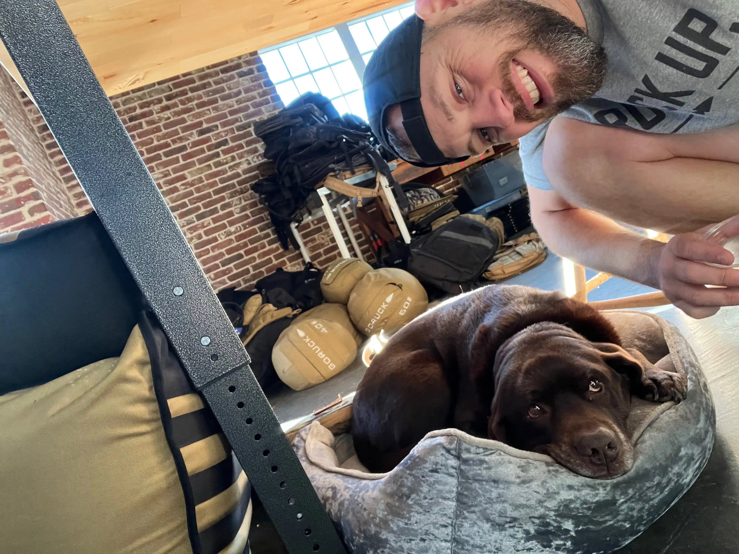 Man in GORUCK shirt smiling next to resting brown dog in bed, rucksack gear in background