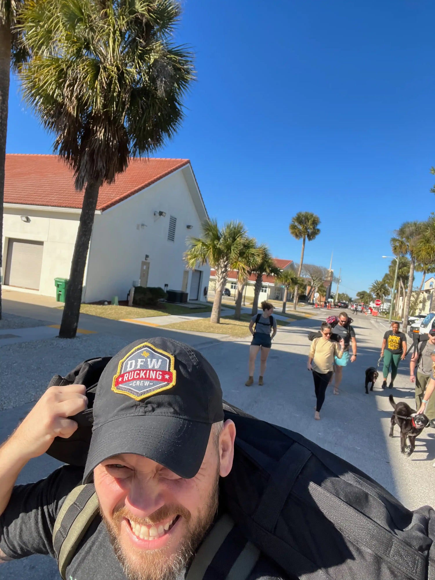 Group rucking outdoors with GORUCK gear under palm trees, sunny day, ruckers and dogs visible.