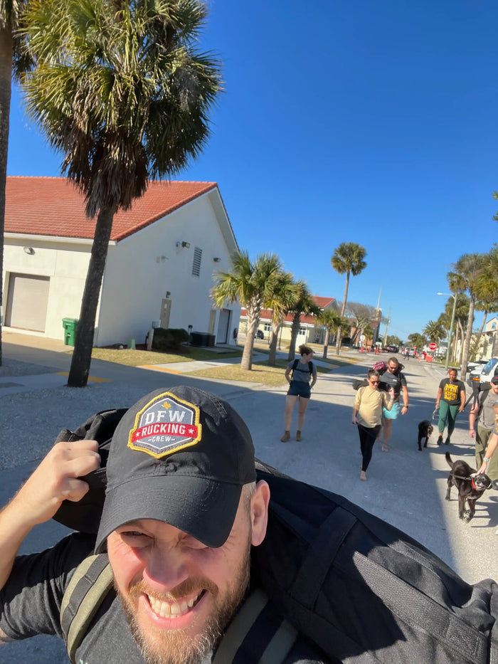 Group rucking outdoors with GORUCK gear under palm trees, sunny day, ruckers and dogs visible.