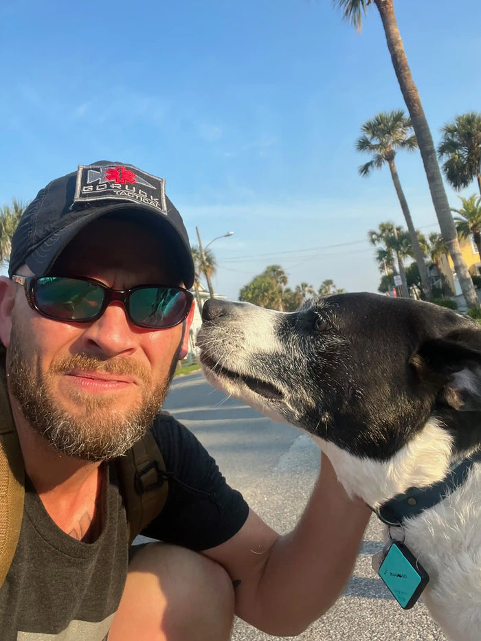 Man wearing GORUCK hat with dog outdoors, palm trees in background, sunny day.
