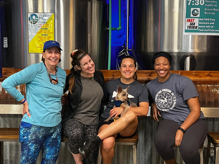 Four women with athletic wear and a small dog at a brewery, GORUCK rucking community event