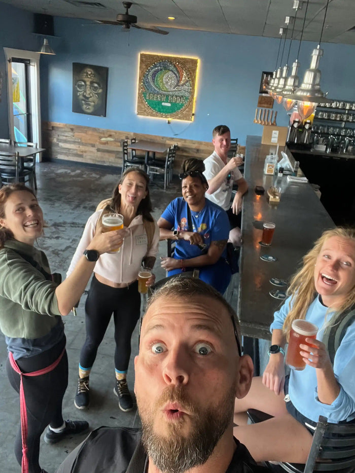 Group of friends in athletic wear celebrating with beers inside a casual bar, goruck.mommunity vibe