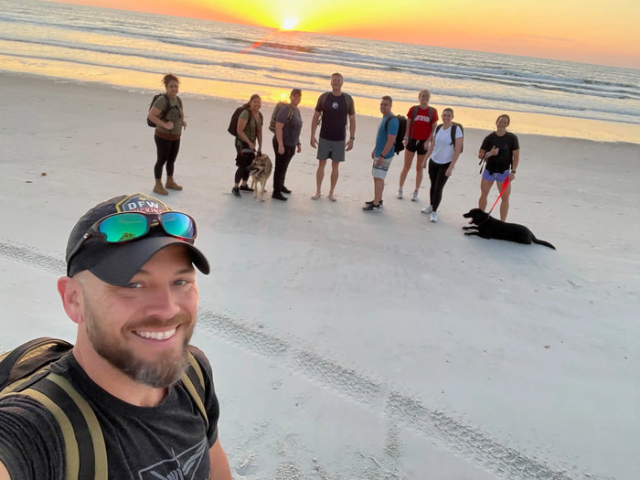 Group of people with backpacks and dogs rucking on the beach at sunset, goruck.mommunity event