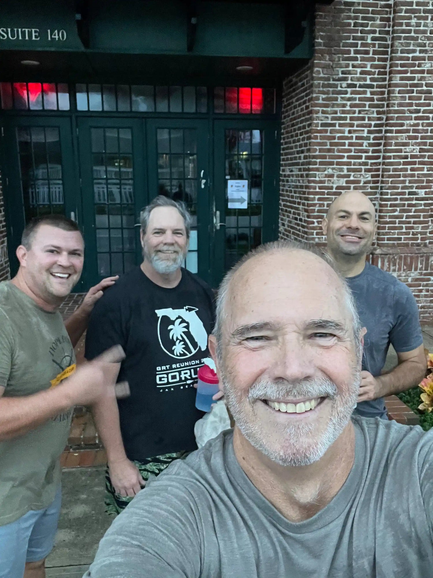 Four smiling men in GORUCK shirts outside brick building, post-rucking event