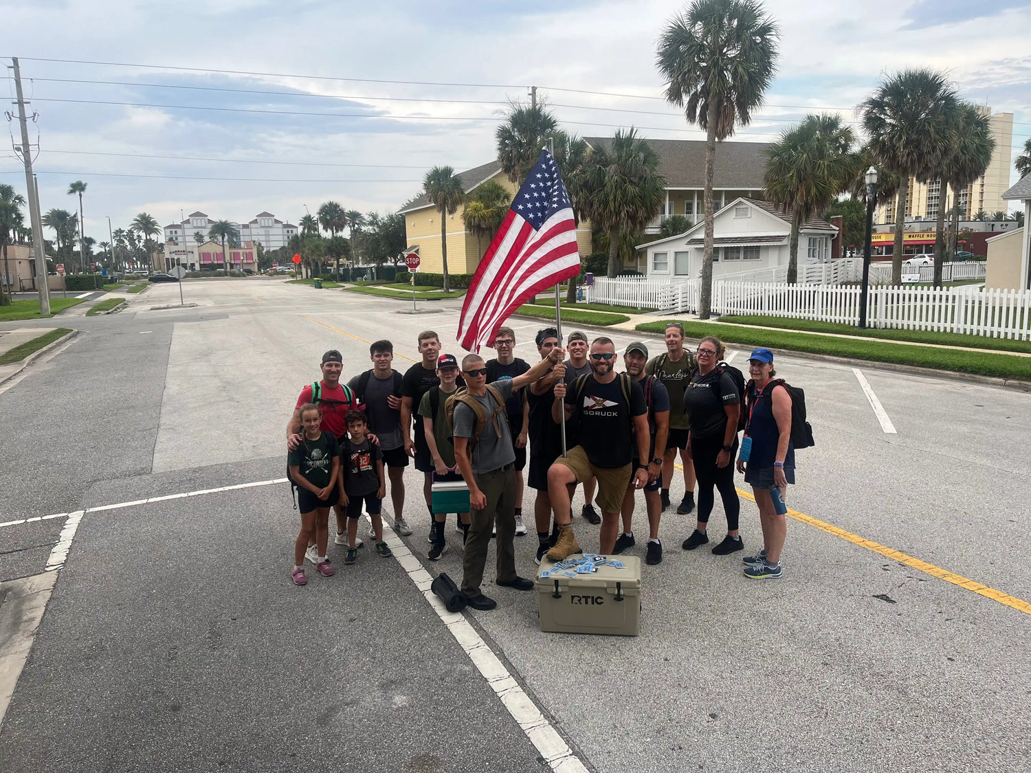 GORUCK group ruck march participants with backpacks and American flag, urban street, palm trees