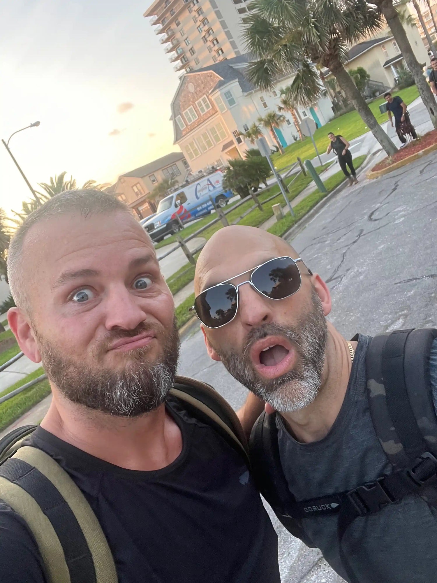 Two men wearing GORUCK backpacks outdoors, making funny faces, with palm trees and houses in the background.