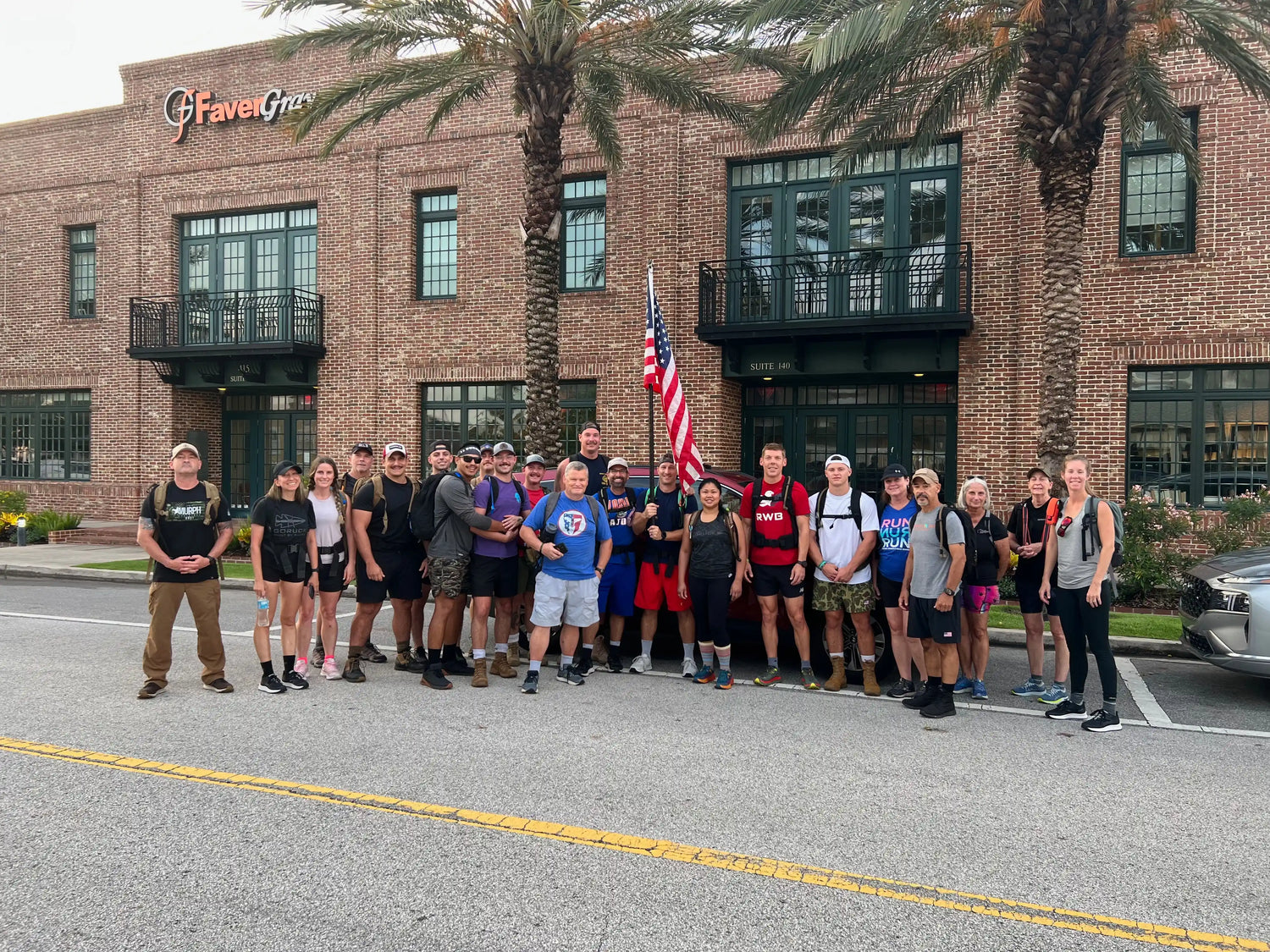 Group of GORUCK participants with rucksacks and American flag outside brick building, palm trees